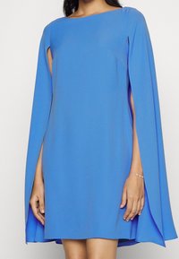 Blue dress with a straight neckline, short length, and cape-style sleeves. Smooth fabric, minimalistic design, and side cut-outs detail.