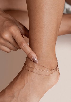 Rose gold layered ankle bracelet with delicate chain links, accentuated by a textured skin tone and a pointed finger highlighting the jewelry.
