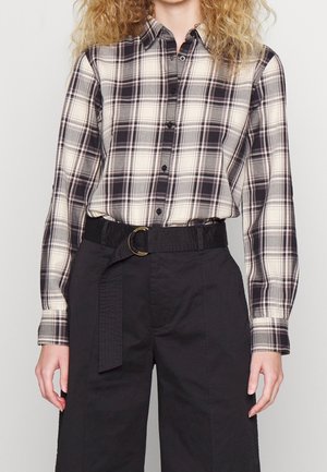 Kvinde iført en sort og hvid ternet skjorte, knappet op og stoppet ned i højtaljede sorte bukser med sort stofbælte og guldlås.