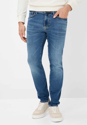Blue denim jeans with a slim fit, featuring five pockets and subtle fading. Paired with light-colored sneakers.