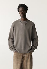 Grauer Pullover aus einem weichen, strukturierten Stoff mit rundem Halsausschnitt und gerippten Bündchen, kombiniert mit beigen Jeans im Denim-Stil, die eine dezente Ausbleichung aufweisen.