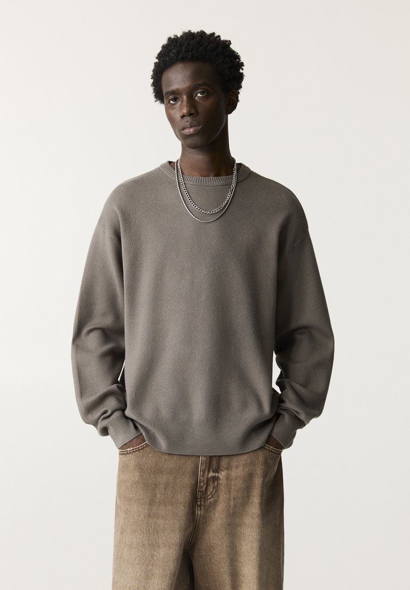Grauer Pullover aus einem weichen, strukturierten Stoff mit rundem Halsausschnitt und gerippten Bündchen, kombiniert mit beigen Jeans im Denim-Stil, die eine dezente Ausbleichung aufweisen.