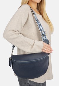 Navy Leder Crossbody-Tasche mit geschwungener Form, ausgestattet mit einem gemusterten blau-beigen Tragegurt und einem Reißverschluss mit Ziehgriff.