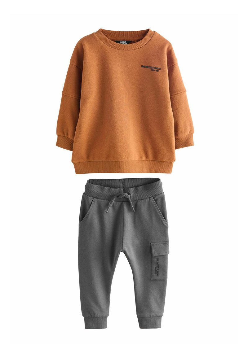 Sweatshirt orange à épaules tombantes avec du texte noir, associé à un pantalon de survêtement gris avec une taille élastique et des poches latérales.