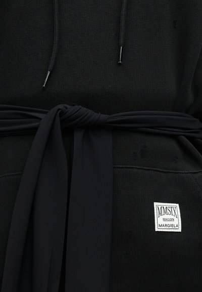 Μαύρο φλις με κορδόνια και δεμένο μαύρο ύφασμα ζώνης γύρω από τη μέση, με λευκή ετικέτα Maison Margiela MMSIX στην τσέπη.