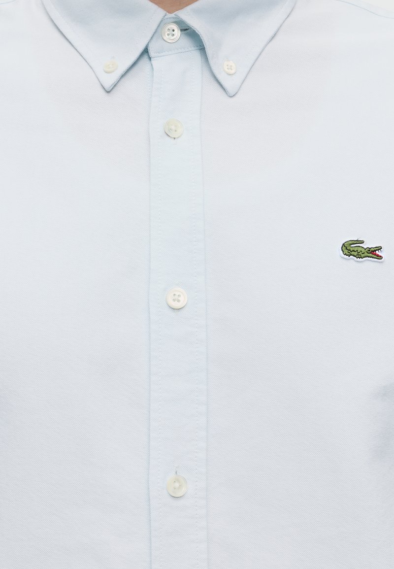 Camicia azzurro chiaro con bottoni, colletto appuntito e piccolo logo verde di coccodrillo sul lato sinistro del petto.