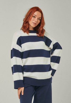 Frau mit roten Haaren, die einen breit gestreiften Pullover in Marineblau und Weiß sowie marineblaue Hosen trägt und mit einer Hand in der Hüfte vor hellem Hintergrund steht.