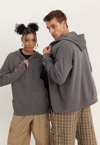 Žena a muž v šedých mikinách s kapucí a béžových kalhotách pózují před jednobarevným bílým pozadím, oba drží límce svých mikin.