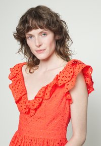Vestido de algodão vermelho com decote em V profundo e mangas com babados, apresentando padrões recortados e um corpete justo que se transforma numa saia godê.