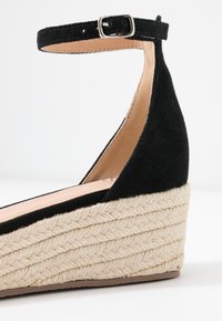 Black suede wedge sandal featuring an ankle strap with a metal buckle, and a textured natural rope sole with a layered design.