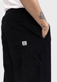 Svarte corduroy-bukser med en teksturert overflate, med en sidelomme og en liten vevd etikett bak. Elastisk midjebånd synlig.