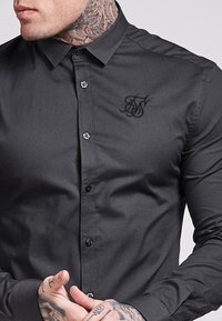 Camisa negra de botones con un diseño ajustado, que presenta un cuello puntiagudo, botones negros y un sutil logo bordado en el pecho.