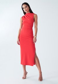 Sarkana, pieguloša kleita ar sānu šķēlumu, teksturētu audumu, bezrokas dizainu un drapētu izgriezumu. Akcentēta ar spīdīgu zelta rokassprādzi.