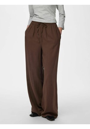 Kvinde iført løse mørkebrune bukser med snor og lommer, kombineret med en lysegrå langærmet top og sorte flade sko.