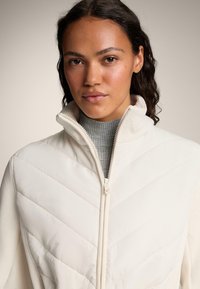 Jachetă albă matlasată cu guler înalt și fermoar, având o textură moale și un model în zigzag; suprapusă peste un pulover turtleneck cu model în dungi de culoare gri deschis.