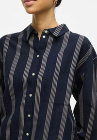 Camisa de botones a rayas marinas con rayas verticales de color claro, un cuello y un bolsillo en el pecho, fabricada con tela texturizada.