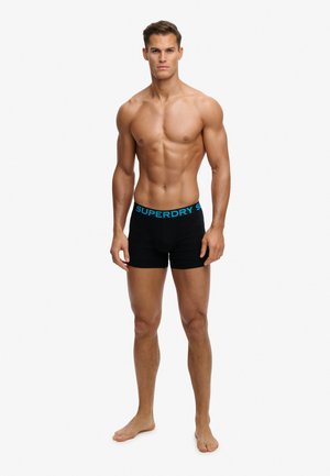 Homme musclé debout pieds nus portant un boxer noir avec une ceinture bleue « SUPERDRY » sur un fond blanc uni.