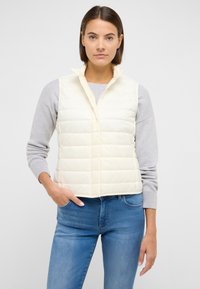 Hellcremefarbene Steppweste mit hohem Kragen, vertikal gestepptem Muster und seitlichen Taschen, kombiniert mit einem grauen Pullover und blauen Jeans.