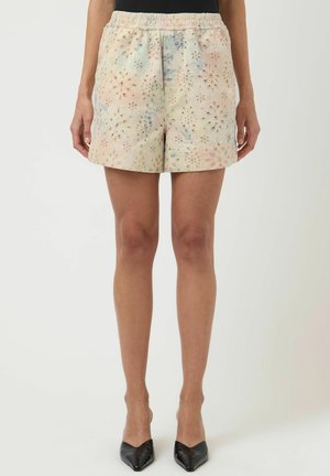 Beige shorts med blomsterprint og elastisk midje, båret av en stående person i svarte spisse hæler.