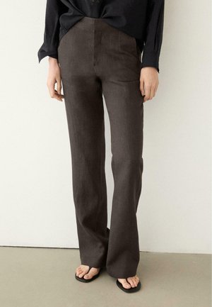 Person iført brune, lige bukser med høj talje, sort langærmet top og sorte sandaler, stående på en lys gulv mod en ensfarvet hvid væg.