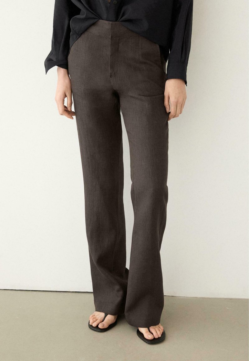 Person iført brune, lige bukser med høj talje, sort langærmet top og sorte sandaler, stående på en lys gulv mod en ensfarvet hvid væg.