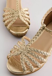 Goldene Riemchensandalen mit geflochtenen Akzenten, offener Zehenpartie, flacher Sohle und einer glatten metallischen Oberfläche auf dem Obermaterial und dem Fußbett.