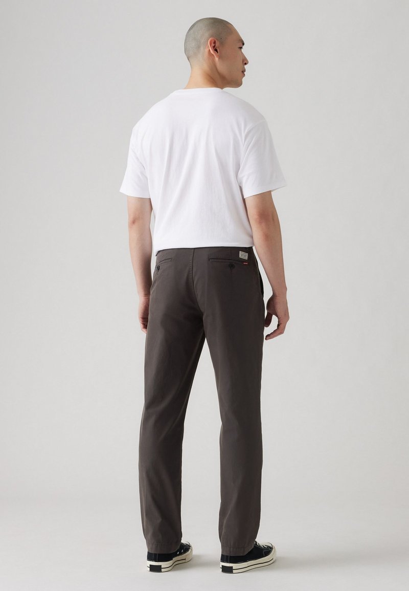 Pantalones grises con un diseño de pierna recta, fabricados en tejido suave, con bolsillos traseros y zapatos negros con suelas blancas. Modelo vistiendo una camiseta blanca.