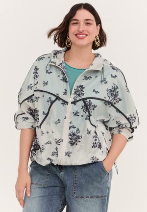 Smilende kvinde med kort mørkt hår bærer guld øreringe i hoop-øreringe, en lys blomsterprintet lynlåsjakke over en teal top og blå jeans med hænderne i lommerne.