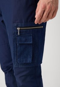Marinebukser har en teksturert denimlomme på siden med glidelås, aksentuert av en liten klafflomme ovenfor. Glatt stoff på den øvre delen.