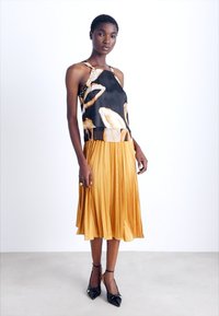 Haut noir avec des motifs crémeux abstraits, associé à une jupe plissée jaune moutarde. Le modèle porte des talons noirs. Tissu lisse, silhouette élégante.