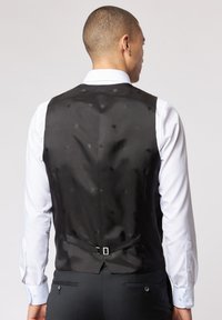 Schwarze Satin-Weste mit dezentem Musterdetail, mit tiefem Rückenausschnitt, verstellbarem Riemen und getragen über einem weißen Hemd.