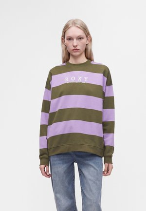 Jeune femme aux longs cheveux blonds porte un pull rayé vert olive et violet avec le texte "ROXY" et un jean bleu, debout devant un fond uni.