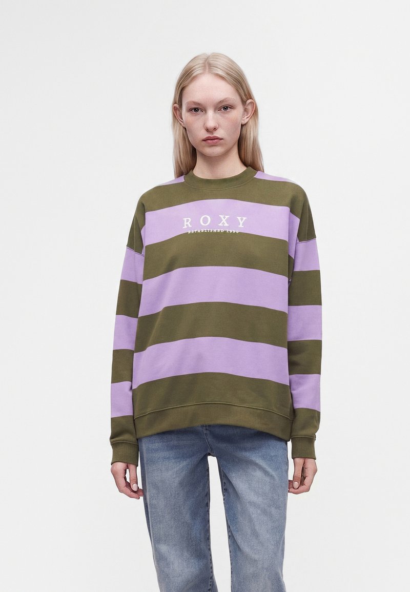 Jeune femme aux longs cheveux blonds porte un pull rayé vert olive et violet avec le texte "ROXY" et un jean bleu, debout devant un fond uni.