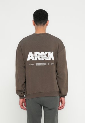 Brązowy oversized sweter z prążkowanymi mankietami i dołem. Posiada dużą białą tekst na plecach. Tkanina wydaje się miękka o matowym wykończeniu.