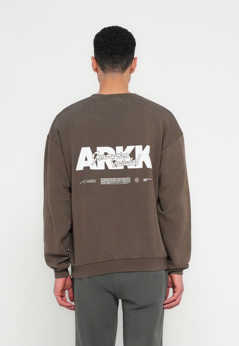 Brown oversized sweatshirt with ribbed cuffs and hem. Features large white text on the back. Fabric appears soft with a matte finish.
