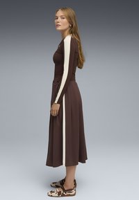 Vestido marrón de mangas largas, con franjas laterales crema y un cintúrón en la cintura. Combinado con zapatos de tela estampados en tonos neutros. Textura de tela suave.