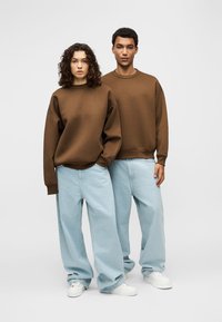 Δύο άτομα που στέκονται δίπλα-δίπλα φορούν καφέ φαρδιές μπλούζες, ανοιχτό μπλε τζιν με φαρδιά πόδια και λευκά αθλητικά σε απλό φόντο.