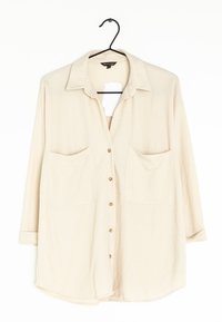 Beige kauluspaita, jossa on kaksi etutaskua ja kolmanneksen mittaiset hihat, valmistettu pehmeästä, tekstuuripinnoitteisesta kankaasta.