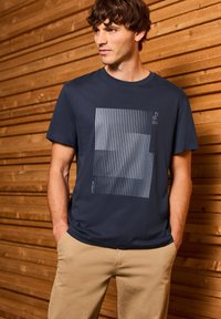 T-shirt en coton bleu marine avec un graphique géométrique gris comportant des rayures verticales, associé à un pantalon beige clair.