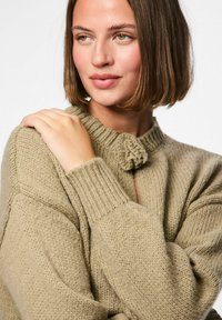 Beige knitted sweater with a round neck and a floral knot detail at the front. Features ribbed cuffs and a chunky texture.