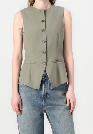 Ärmellose olivgrüne Weste mit Knöpfen, kombiniert mit blauen Denim-Jeans, zeigt ein Tattoo am linken Oberarm.