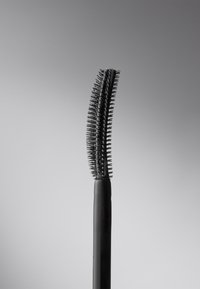 Scovolino per mascara curvo con manico in gomma nera; presenta setole corte e dense da un lato e setole più lunghe dal lato opposto.