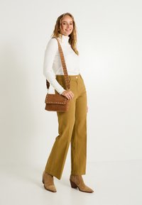 Bolso de ante marrón claro con correa decorada con tachuelas, combinado con pantalones de campana mostaza y un suéter de cuello alto blanco. El conjunto presenta un ajuste relajado.