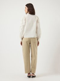Vrouw die met de rug naar ons staat, gekleed in een witte blouse met pofmouwen en beige wijde broek met zwarte haksandalen.