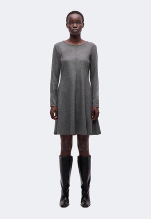 Femme debout face caméra, portant une robe grise à manches longues et des bottes noires montant jusqu’aux genoux, sur un fond uni.