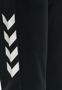 Zwarte sportbroeken met een gladde textuur en een wit chevronpatroon aan de zijkant. Het ontwerp richt zich op comfort en rekbaarheid.