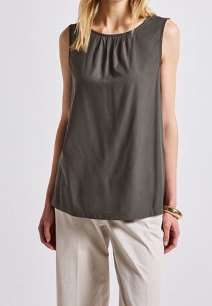 Femme portant un haut sans manches vert olive foncé avec un col rond et un pantalon beige clair, accessoirisée d'un bracelet doré au poignet.