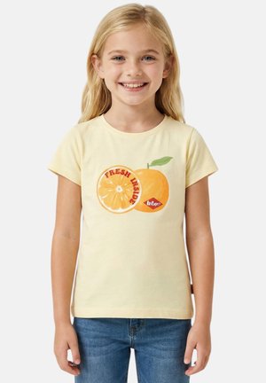 Fille souriante aux longs cheveux blonds portant un t-shirt jaune avec un motif orange et le texte « Fresh Inside » et un jean bleu.