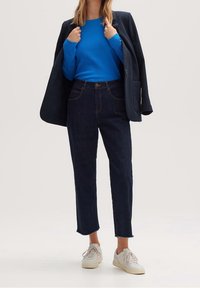 Blauwe kabeltrui, donkere jeans met rafelige zoom en witte sneakers, aangevuld met een zwarte blazer. Casual, gestructureerde outfit voor verschillende gelegenheden.