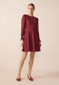 Robe bordeaux avec de longues manches transparentes, un corsage ajusté et une jupe évasée. Portée avec des escarpins assortis à bout pointu ornés d'un noeud.